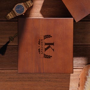 Peut inclure: Une bo&icirc;te en bois avec le nom "KRISTIAN" et la lettre "K" grav&eacute;s sur le couvercle. Une montre dor&eacute;e avec un cadran bleu et une cl&eacute; avec un pompon noir sont &eacute;galement visibles. Les objets sont sur une surface en bois.