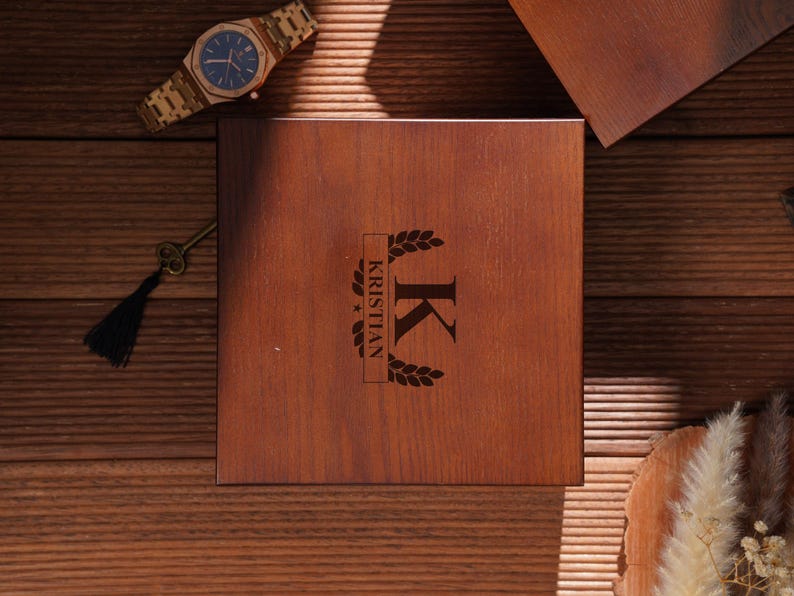 Peut inclure: Une bo&icirc;te en bois avec le nom "KRISTIAN" et la lettre "K" grav&eacute;s sur le couvercle. Une montre dor&eacute;e avec un cadran bleu et une cl&eacute; avec un pompon noir sont &eacute;galement visibles. Les objets sont sur une surface en bois.