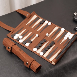 Könnte beinhalten: Ein braunes Backgammon-Set mit schwarzen und weißen Spielsteinen, Würfeln und einer Flasche. Das Spielbrett ist geöffnet und zeigt die Spielfläche mit einem Reißverschluss. Ein aufgerolltes Etui befindet sich neben dem Spielbrett.
