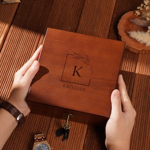 Peut inclure: Une bo&icirc;te en bois avec le nom "Kristian" grav&eacute; sur le couvercle, ainsi que la lettre "K" dans un carr&eacute; avec un motif de feuilles. Une montre et une cl&eacute; dor&eacute;es sont &agrave; c&ocirc;t&eacute; de la bo&icirc;te.