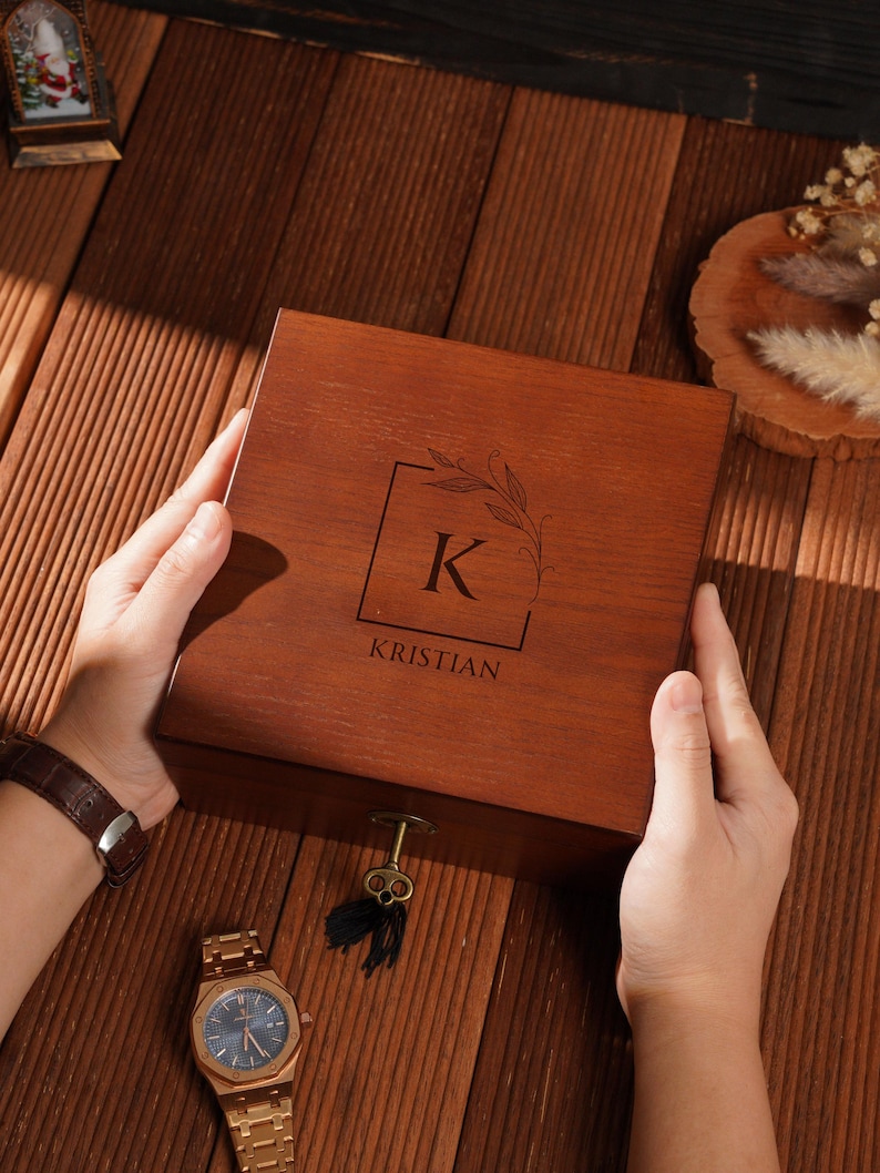Peut inclure: Une bo&icirc;te en bois avec le nom "Kristian" grav&eacute; sur le couvercle, ainsi que la lettre "K" dans un carr&eacute; avec un motif de feuilles. Une montre et une cl&eacute; dor&eacute;es sont &agrave; c&ocirc;t&eacute; de la bo&icirc;te.