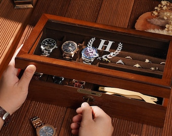 May include: A wooden watch and jewelry box with a glass top, showcasing several watches and rings. The box is open, revealing a drawer with a key. The watches have gold and silver tones. The box is on a wooden surface.