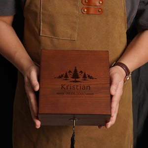 Peut inclure: Une bo&icirc;te en bois fonc&eacute; avec un motif de montagne et d'arbres est tenue par une personne portant un tablier marron. La bo&icirc;te porte le nom "Kristian" et la date "08.08.2000". Une cl&eacute; avec un pompon noir est suspendue.