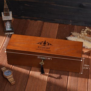 Peut inclure: Un coffret rectangulaire en bois avec une serrure et un pompon, mesurant 35,5 cm de long et 10 cm de haut. La bo&icirc;te est grav&eacute;e d'un motif de montagne et d'arbres, ainsi que du nom "Kristian" et de la date "08.08.2000".