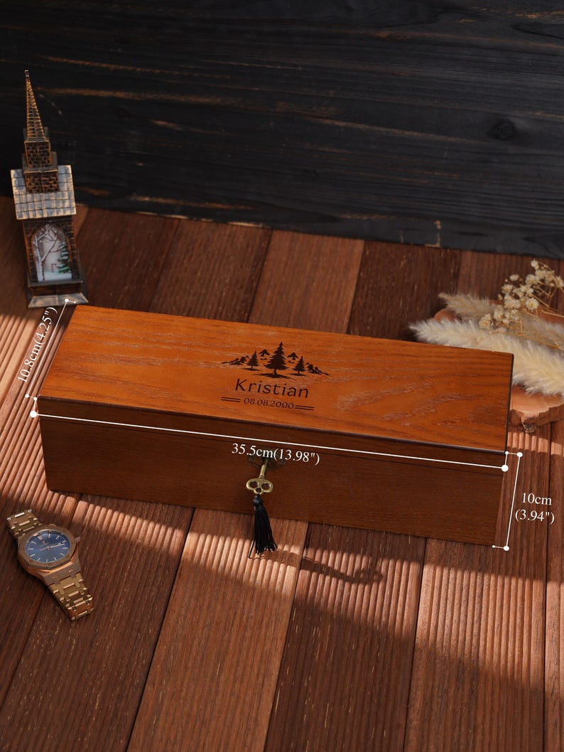 Peut inclure: Un coffret rectangulaire en bois avec une serrure et un pompon, mesurant 35,5 cm de long et 10 cm de haut. La bo&icirc;te est grav&eacute;e d'un motif de montagne et d'arbres, ainsi que du nom "Kristian" et de la date "08.08.2000".