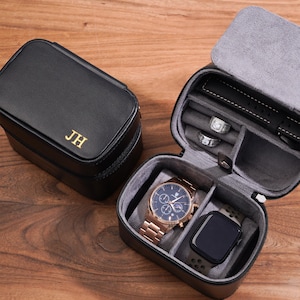 Könnte beinhalten: Schwarzes Leder-Uhrenetui mit goldenen Initialen "JH". Das offene Etui zeigt eine roségoldene Uhr, eine schwarze Smartwatch und zwei Ringe. Der Innenraum ist mit grauem Stoff ausgekleidet, und das Etui befindet sich auf einer Holzoberfläche.