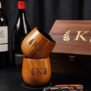 May include: Two wooden cups, each engraved with the letter "K" and a leaf design, are stacked on a black surface. A wooden box with the letter "K" and a leaf design is in the background. A bottle opener and a wine bottle are also visible.