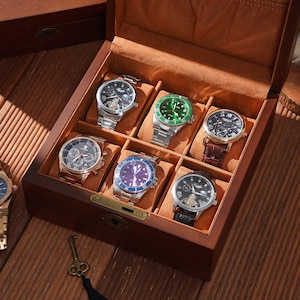 Peut inclure: Une bo&icirc;te &agrave; montres en bois avec huit compartiments, chacun contenant une montre diff&eacute;rente. Les montres ont diff&eacute;rentes couleurs de bracelet et de cadran, dont l'argent, le noir, le vert et le bleu. Une cl&eacute; avec un pompon noir est pos&eacute;e sur la surface en bois.