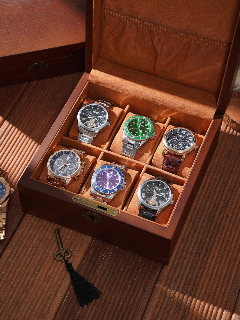 Peut inclure: Une bo&icirc;te &agrave; montres en bois avec huit compartiments, chacun contenant une montre diff&eacute;rente. Les montres ont diff&eacute;rentes couleurs de bracelet et de cadran, dont l'argent, le noir, le vert et le bleu. Une cl&eacute; avec un pompon noir est pos&eacute;e sur la surface en bois.