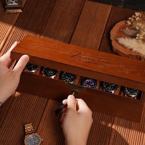 Peut inclure: Un coffret en bois fonc&eacute; pour montres avec un couvercle &agrave; charni&egrave;re, contenant plusieurs montres-bracelets. La bo&icirc;te est ouverte avec une cl&eacute;. Une montre-bracelet dor&eacute;e est visible sur la surface en bois. La bo&icirc;te est grav&eacute;e d'un nom et d'une date.