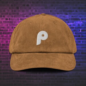 Può includere: Un cappellino da baseball in velluto a coste marrone con una lettera P ricamata in bianco sul pannello frontale. La lettera P include un piccolo dettaglio a forma di stella marrone ritagliata. Il cappellino ha una corona strutturata e visiera curva.