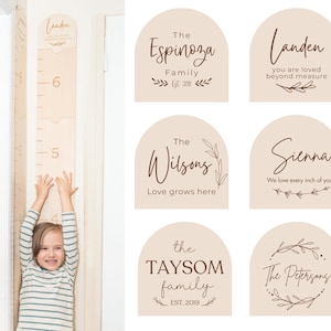 May include: A wooden growth chart with inch markings and the name "Landen" at the top. A child is standing next to the chart, arms raised. Also included are six beige arched signs with family names and phrases in brown script.