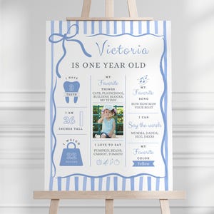 May include: A light blue and white striped milestone birthday sign featuring the text Victoria Is One Year Old, with sections for teeth, height in inches, weight in pounds, favorite things, song, words, and color, plus a central photo placeholder.