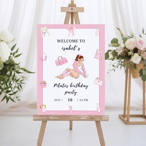 May include: A pink Pilates birthday party sign on a wooden easel. The sign features the text "Welcome to Isabel's" and "Pilates birthday party" with the date and time. The sign is decorated with illustrations of Pilates-related items.