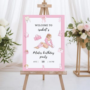May include: A pink and white Pilates birthday party sign on a wooden easel. The sign features illustrations of workout gear and a woman in workout clothes. Text reads "Welcome to Isabel's Pilates birthday party" with the date and time.