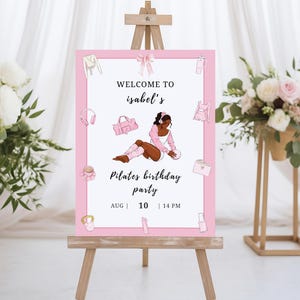 May include: A pink Pilates birthday party sign on a wooden easel. The sign features the text "Welcome to Isabel's" and "Pilates birthday party" with the date and time. The sign is decorated with illustrations of workout gear.