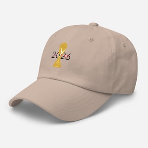 Puede incluir: Gorra de béisbol beige con un diseño de trofeo dorado y el texto "2026". La gorra tiene una visera curva y un botón en la parte superior. El diseño está en la parte delantera de la gorra.