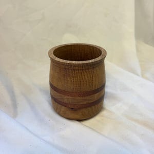 May include: A cylindrical wooden cup featuring a light brown grain pattern with two distinct horizontal dark reddish-brown bands encircling the middle section. The cup has a smooth, rounded rim and a slightly tapered base, showcasing natural wood textures.