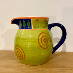 May include: A decorative ceramic jug with a green body and a blue handle. The jug features red spiral designs and floral accents. The top rim has blue and orange geometric patterns. A unique piece for serving beverages.