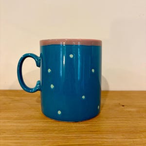 May include: A teal ceramic mug with a rounded handle and a pink rim. The mug is decorated with small, evenly spaced white polka dots. The mug is sitting on a wooden surface.