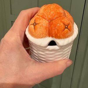 May include: A ceramic butter dish in orange and white. The top is orange with black detailing, and the base is white with a textured pattern. The dish is held in a hand.