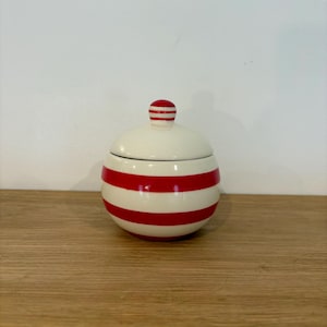 May include: A cream-coloured ceramic container with a lid, featuring bold red horizontal stripes. The lid is cream with red stripes. The container sits on a wooden surface. A decorative item, possibly for storage.