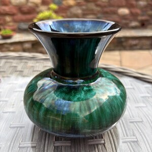May include: A small ceramic vase with a wide, flared rim and a rounded, bulbous body. The glaze features a deep, glossy emerald green finish with swirling patterns of dark blue and black, creating a reflective, multi-tonal appearance.