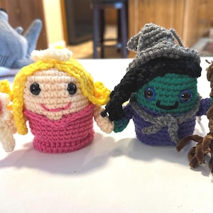 May include: Two handmade crochet dolls: a princess with blonde hair and a pink dress, and a green-skinned witch with a black hat and broom. The dolls are made with yarn and have large, expressive eyes. They are standing side-by-side.