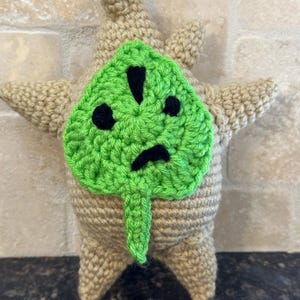 Könnte beinhalten: Handgemachte Häkelpuppe in Form eines Korok-Charakters. Die Puppe ist hauptsächlich beige mit einem grünen Blattgesicht. Das Blatt hat schwarze Augen und einen Mund. Die Puppe hat ausgestreckte Arme und einen spitzen Kopf.