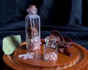May include: Two clear glass bottles containing small pink mineral crystals. The taller bottle is sealed with a cork and topped with pink wax drips, containing dried botanical elements. The shorter bottle is open, filled with pink crystals.