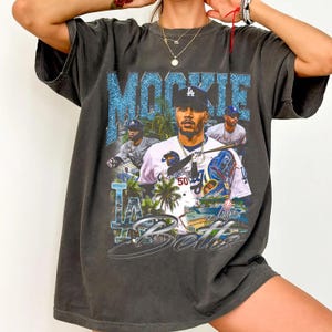 May include: Dark gray t-shirt featuring a graphic of Mookie Betts in a Dodgers uniform. The design includes the text "Mookie Betts" in a blue glitter font, along with images of him playing baseball and the Dodgers logo. The shirt has a vintage, worn look.