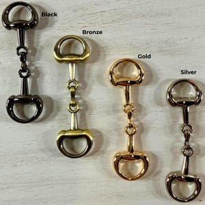 May include: Four metal horse bit hardware pieces arranged vertically, labeled Black, Bronze, Gold, and Silver. Each piece features a central bar with two circular loops at the ends, connected by a small central link, showcasing distinct metallic finishes.