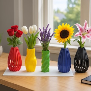May include: A collection of colorful, 3D-printed vases, each holding a different type of flower. The vases are red, yellow, green, blue, and black. Flowers include roses, tulips, lavender, a sunflower, and a pink lily. The vases are on a wooden desk.