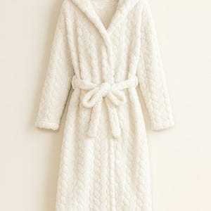 Pode incluir: Um roupão branco com capuz, com um padrão texturizado e um cinto a condizer amarrado em laço. O roupão está pendurado num cabide de madeira sobre um fundo branco liso. As mangas compridas e o design até ao joelho sugerem conforto.