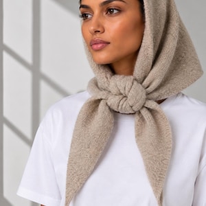 May include: A beige knitted headscarf tied in a knot under the chin, featuring a soft, textured fabric construction. The headscarf is worn over a plain white t-shirt, covering the top of the head and draping down to the chest in a triangular shape.