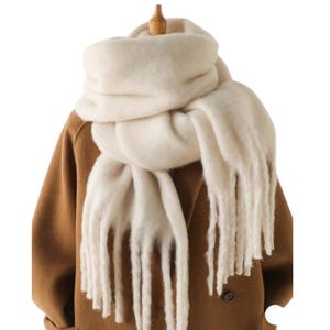 May include: Cream-colored, oversized scarf with long fringe. The scarf is draped over a brown coat, creating a cozy and stylish look. The soft, fluffy texture suggests warmth and comfort, perfect for cold weather.