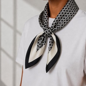 May include: A square silk scarf tied around the neck, featuring a black and white geometric chain-link pattern. The scarf has a solid black border and a cream-colored interior section, worn over a plain white t-shirt with a simple crew neckline.