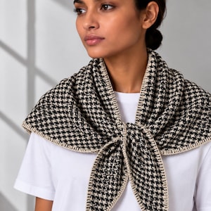 May include: A triangular scarf featuring a black and white houndstooth pattern with a solid beige crocheted border. The scarf is tied in a knot at the front and is worn over a plain white short-sleeved t-shirt with a simple crew neckline.