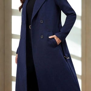 Puede incluir: Un abrigo azul marino de doble botonadura con cinturón y bolsillos. El abrigo largo tiene un diseño clásico y es adecuado para diversas ocasiones. El abrigo presenta el texto "RAVISHA CREATION" en un logotipo en la esquina superior derecha.