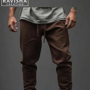May include: Brown drawstring trousers with matching boots. The trousers have a relaxed fit and are gathered at the ankles. The boots are a similar shade of brown and appear to be made of a soft material. The top is a grey t-shirt. The logo "RAVISHA CREATION" is in the top left corner.
