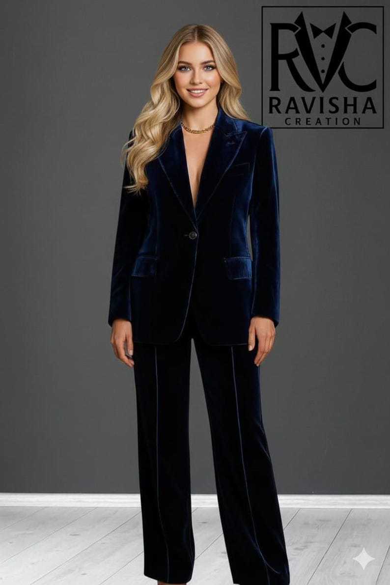 May include: A navy blue velvet suit, consisting of a blazer and matching trousers. The blazer has a single-button closure and notched lapels. The trousers have a straight leg. The logo "RAVISHA CREATION" is visible in the upper right corner.