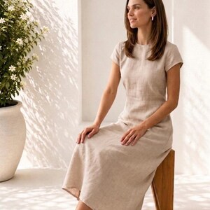 May include: A beige midi-length dress with short sleeves, a round neckline, and a fitted bodice that flows into a relaxed skirt. The garment is made of a textured fabric and is paired with beige slip-on shoes. The design is simple and neutral.