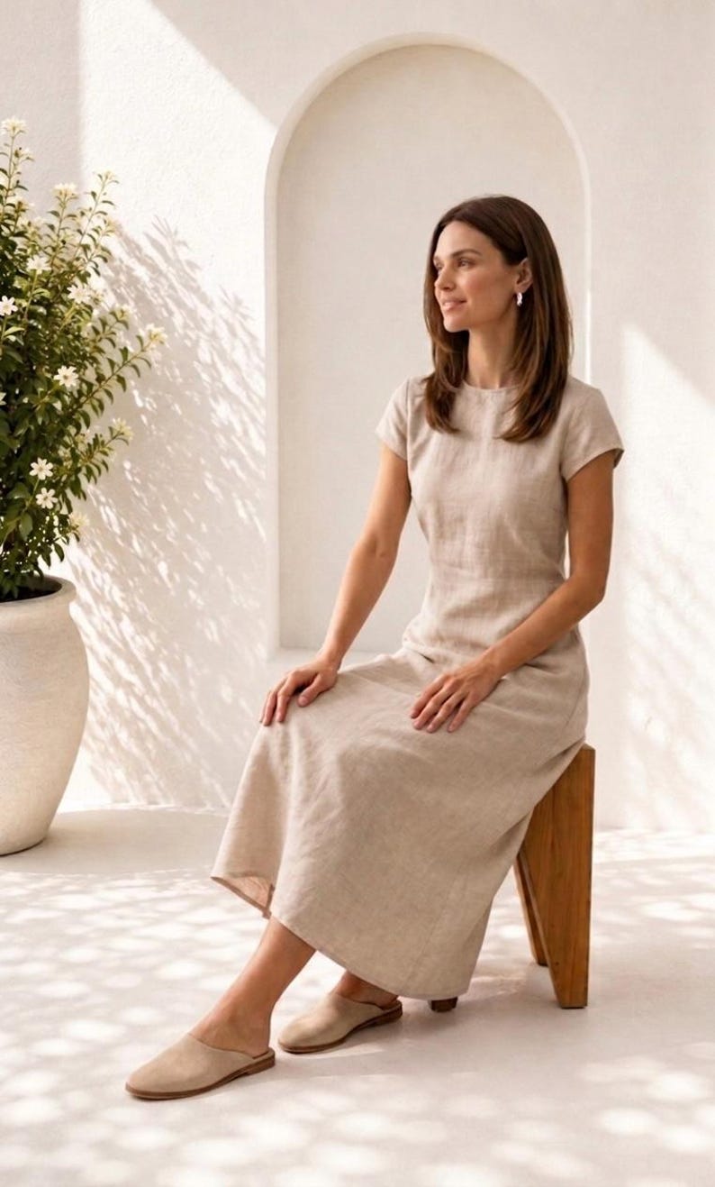 May include: A beige midi-length dress with short sleeves, a round neckline, and a fitted bodice that flows into a relaxed skirt. The garment is made of a textured fabric and is paired with beige slip-on shoes. The design is simple and neutral.