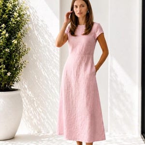 May include: A light pink midi-length dress with short sleeves, a round neckline, and a fitted waistline. The dress features a simple, solid-colored design with a straight skirt that falls below the knees, paired with neutral-toned slip-on shoes.
