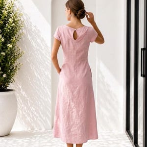 May include: A light pink midi-length dress with short sleeves, a round neckline, and a teardrop-shaped cutout detail at the upper back featuring a single button closure. The garment has a fitted bodice and a flared skirt with side pockets.