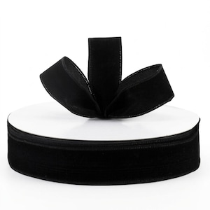 Puede incluir: Un carrete de cinta de terciopelo negro sólido con una superficie suave y texturizada. La cinta está enrollada firmemente alrededor de un núcleo de cartón blanco circular, con un pequeño lazo de la misma cinta negra hacia arriba.