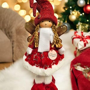 May include: A Christmas fairy doll with a red knitted hat and dress, gold wings, and a white scarf. The doll has long blonde braids and is holding a tag that says "Michelle's Exclusive". The doll is on a white surface.