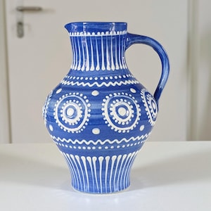 Può includere: Brocca in ceramica blu con manico, decorata con linee verticali bianche e motivi circolari. La brocca ha un corpo arrotondato e un beccuccio. Un elemento decorativo per la casa.