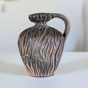 May include: A small, textured ceramic jug with a handle. The jug is primarily grey with brown and pink accents. The surface has a carved, vertical, wavy pattern. The jug is sitting on a white surface.