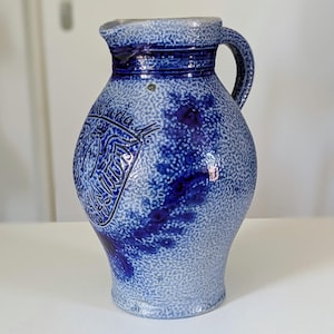 Può includere: Una brocca in gres smaltato al sale con una superficie testurizzata in blu chiaro e grigio maculato, dotata di manico e bordo blu scuro. La parte anteriore presenta uno stemma in rilievo blu scuro con un'aquila e motivi floreali.